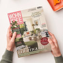 Wohnmagazin „WOHN IDEE“ in schräger Draufsicht, von zwei Händen gehalten, auf weißer Fläche mit orangefarbenem Gegenstand oben rechts. Sichtbare Titel: „Garten-Checkliste – Was Beete und Pflanzen jetzt brauchen“, „Die pure Leichtigkeit“ sowie Hinweise zu Pflanzen, Ordnung, Licht, Feng-Shui, „Bitte zu Tisch“ und „Der große Sofa-Guide“.