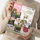 Eine Person im beigefarbenen Strickpullover hält eine schräg fotografierte Wohnzeitschrift mit rosafarbener Titelseite „WOHN IDEE – Für ein Zuhause!“. Sichtbare Texte: „Garten-Checkliste“, „Bitter zu Tisch“, „Kühle Brise“, „Der große Sofa-Guide“, „Die pure LEICHTIGKEIT“ und „Sommer-Gewinnspiel Heiße Preise“; das Titelbild zeigt ein helles Wohnzimmer mit grünen Sofas, Pflanzen und Couchtisch.