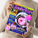 Weibliche Person im beigefarbenen Strickpullover hält ein schräg fotografiertes Magazin mit blau-gelbem Cover. Sichtbare Texte: „Zaubertopf“, „DAS SAGT DIR KEINER: 10 geheime Mix-Tipps“, „Lieblingsrezepte für Thermomix®“, „5 Zutaten“, „Nudelsalate“ und „Lust auf Eis mit Thermomix!“; abgebildet sind ein Thermomix mit rosa Eiscreme, Erdbeeren, Tomaten und eine blonde Frau.