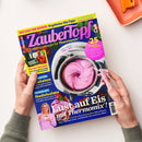 Schräg von oben fotografierte Zeitschrift „ZauberTopf“ mit dunkelblauem, gelbem und pinkem Cover, gehalten von zwei Händen über weißer Fläche; darauf eine Thermomix-Schüssel mit pinker Eismasse, Erdbeeren sowie eine Frau mit Dessert. Sichtbare Texte: „Lieblingsrezepte für Thermomix“, „Alle Rezepte auch für TM7“, „+25 Express-Rezepte“, „Nudelsalate“ und „Lust auf Eis mit Thermomix!“. Oben rechts liegt ein orangefarbenes Gerät.
