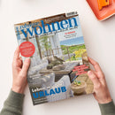 Schräg auf einem weißen Tisch gehaltene Wohnzeitschrift mit zwei Händen und grauen Strickärmeln. Das Cover zeigt eine helle Terrasse mit Sitzmöbeln, Holztisch, Pflanzen und Meerblick sowie die Texte „neue zuhause Wohnen“, „Wie Wohnen wirklich glücklich macht“, „12 Regale“, „Genuss weltweit“, „Blickfang Bilder“, „Kaffee & Eis“ und „Leben wie im Urlaub“.